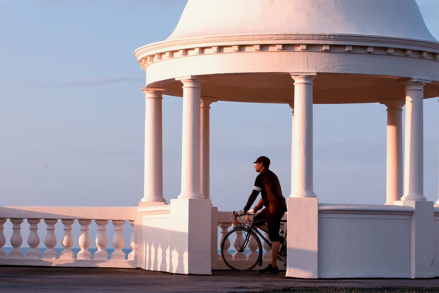 Bicycles in the Landscape: Cyclist and photographer Roff Smith poses with his touring bicycle inside the Tuscan columned dome of the King George V Colonnade at Bexhill on Sea. The Grade II listed monument was built in 1911 to commemorate the coronation of King George V. Its neoclassical lines, balustrades, dome and Tuscan columns and the Union Jack seemed a perfect setting for a series of images celebrating the beauty of a British-built classic touring bicycle