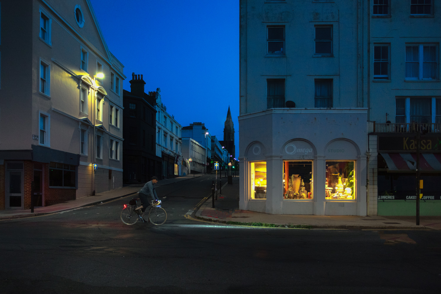 Nighthawk - A Bouquet to Edward Hopper. A cyclist pedals past a dark urban street corner l