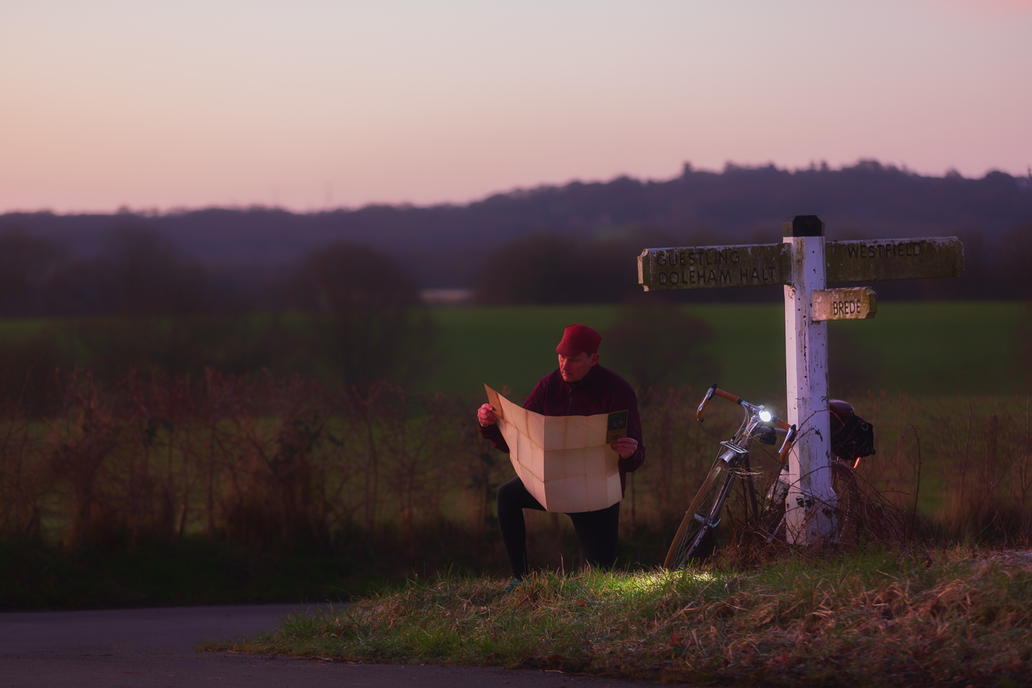 Cyclist consulting a map by lamplight at a lonely crossroads at dusk